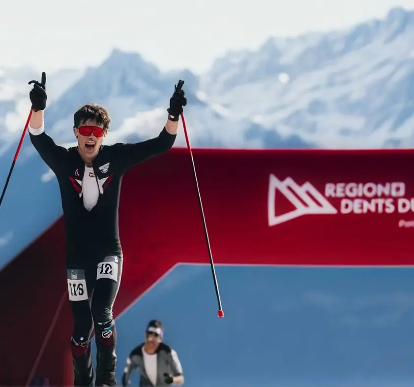 Man in a suit and goggles, holding ski poles in his hands, standing atop a snow-covered slope