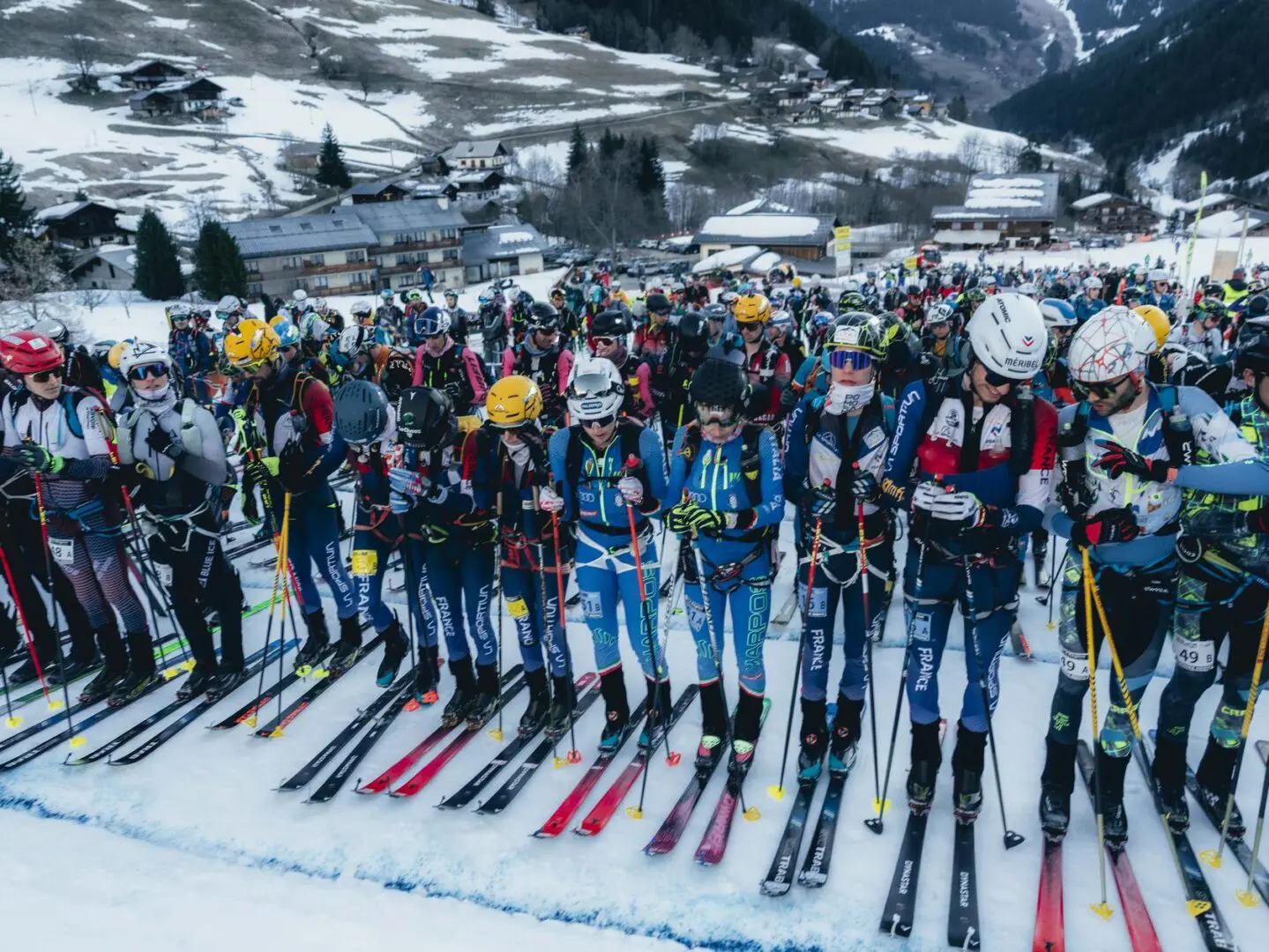 Group of people standing on top of a snow covered slope, all wearing helmets, goggles, gloves, and holding ski poles