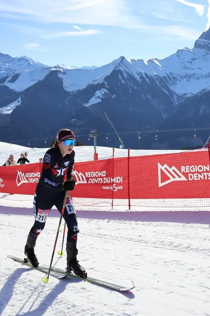 Woman wearing goggles, a cap, gloves, and shoes, holding ski poles and standing on skis atop a snow-covered slope