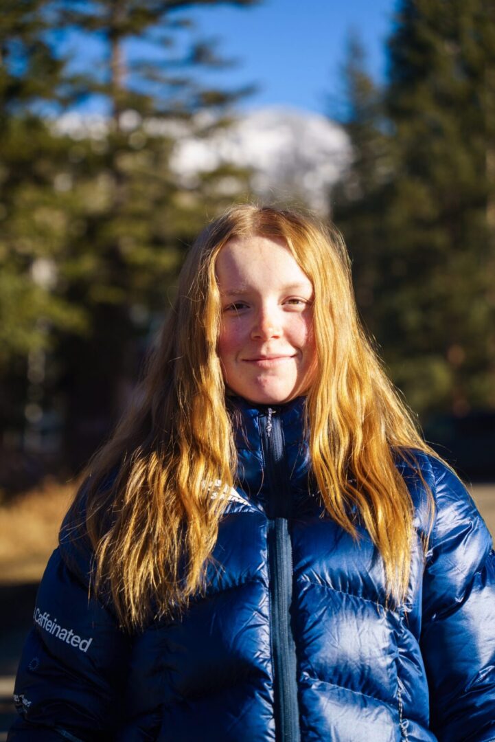 Girl in a jacket standing in front of a backdrop of trees and a clear blue sky