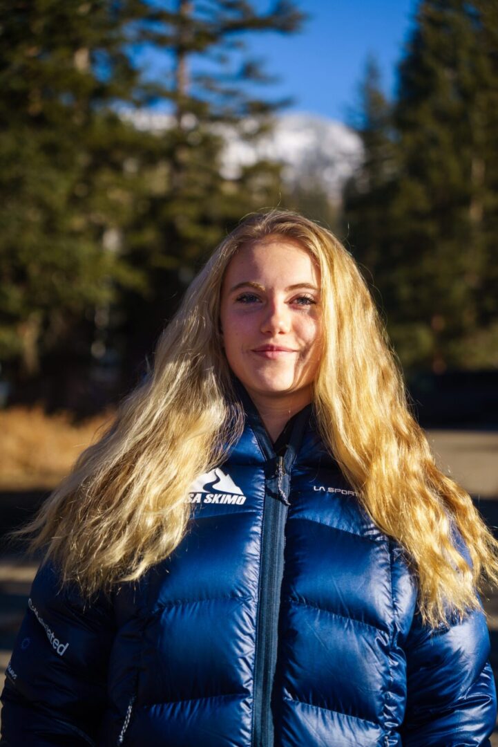 Woman in a jacket standing in front of a backdrop of trees, mountains, and a clear blue sky