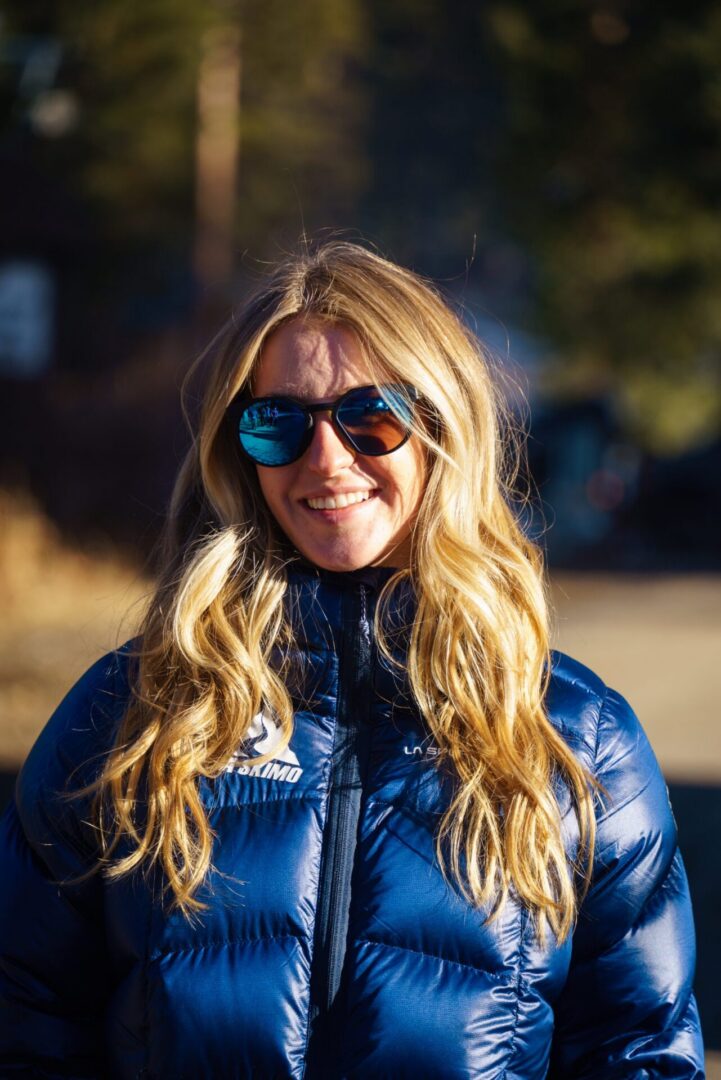 Woman in a jacket and sunglasses standing in front of a backdrop of trees