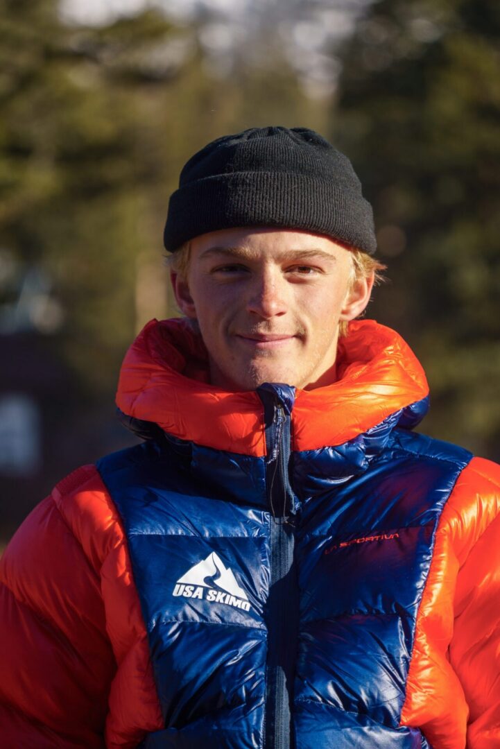 Man wearing a blue and jacket and a black beanie, standing in front of a backdrop of trees
