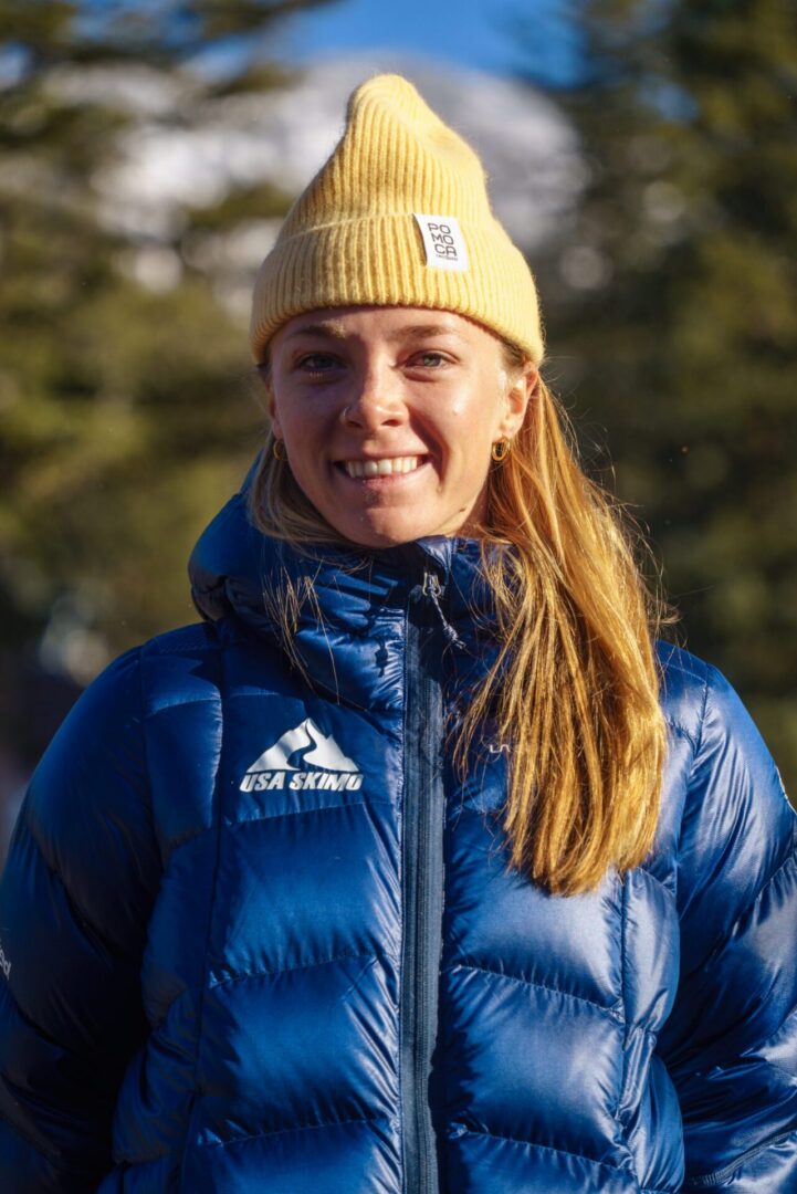 Woman in a jacket and hat standing in front of a backdrop of trees and a clear blue sky