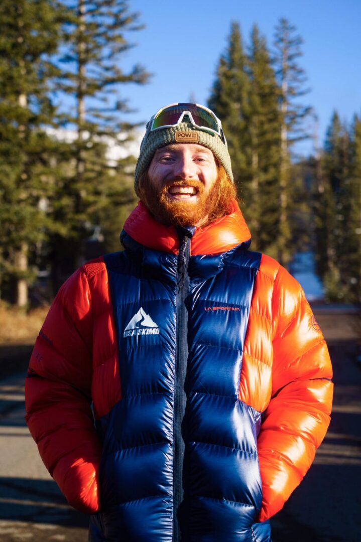 Man with a beard wearing a red and jacket and a cap, at the camera